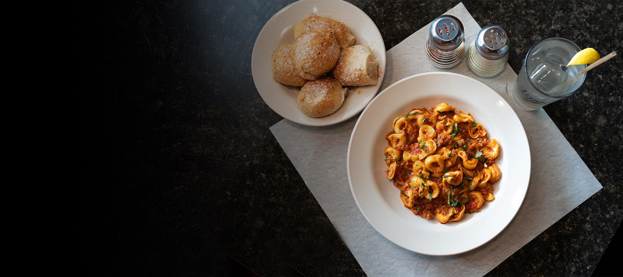 Cheese tortellini bolognese in a white bowl sitting on a table next to a bowl of bread rolls and water glass